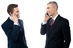 Two businessmen talking on the phone and looking at each other.