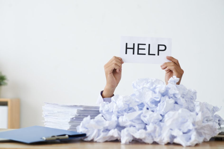 Person under heap of crumpled papers with hand holding a help sign.