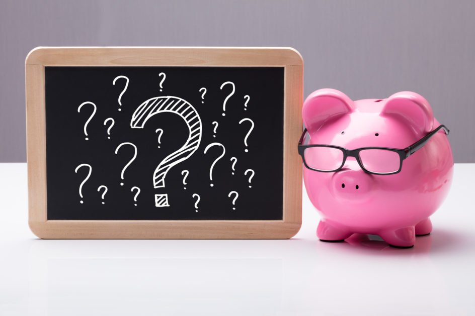 Close-up Of Piggy Bank With Eyeglass And Blackboard With Question Marks On Desk